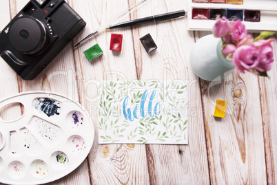 Artist workspace. Hello written in calligraphy style, palette, watercolor, brush, vintage photo camera, bouquet of roses on a wooden background.