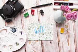 Artist workspace. Hello written in calligraphy style, palette, watercolor, brush, vintage photo camera, bouquet of roses on a wooden background.
