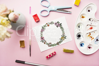 Artist workspace. Floral wreath frame hand painted with watercolor, bouquet of chrysanthemum and roses, glasses, paintbrush, scissors, watercolor, palette on a pink background.