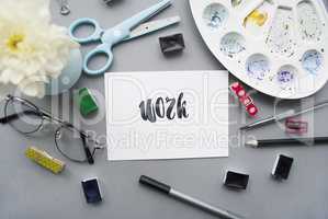 Artist workspace. Word work written in calligraphy style, chrysanthemum, glasses, paintbrush, scissors, watercolor, palette on a gray background. Flat lay
