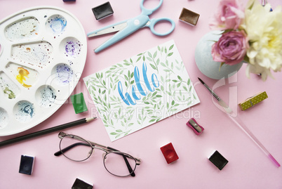 Artist workspace. Hello written in calligraphy style, palette, watercolor, brush, glasses, scissors, bouquet of flowers on a pink background. Flat lay