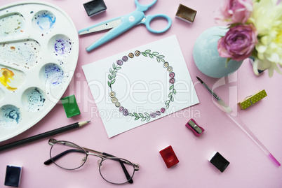 Artist's workspace. Floral wreath frame painted with watercolor, vase with a bouquet of flowers, glasses, paintbrush, scissors,