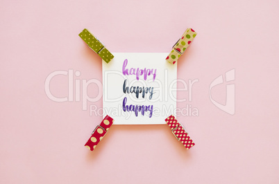 Happy handwritten with watercolor in calligraphy style, miniature clothespins on a pink background. Flat lay, top view