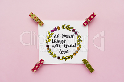 Inspirational quote do small things with great love handwritten with watercolor in calligraphy style, miniature clothespins on a pink background. Flat lay