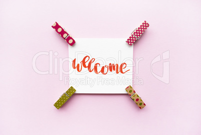 Word Welcome handwritten with watercolor in calligraphy style, miniature clothespins on a pink background. Flat lay