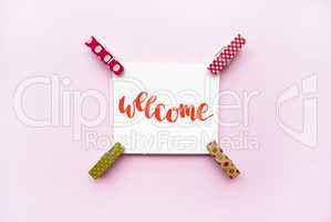 Word Welcome handwritten with watercolor in calligraphy style, miniature clothespins on a pink background. Flat lay