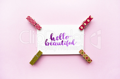Inspirational quote Hello beautiful handwritten with watercolor in calligraphy style, miniature clothespins on a pink background. Flat lay