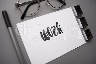 Composition with word work written in calligraphy style, liner, black watercolor, glasses on gray background. Top view