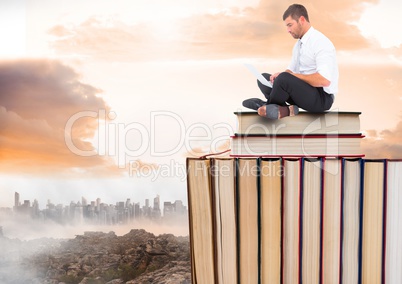 Man with laptop sitting on Books stacked by distant city and clouds