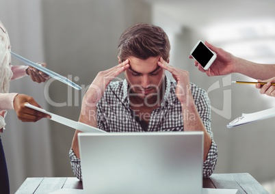 Stressed man on laptop desk surrounded by files phones tablets in office