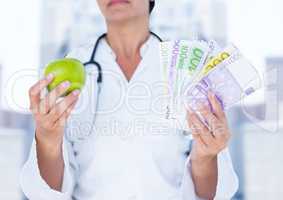 Doctor mid section with apple and money against blurry buildings