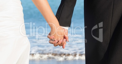 Bride and groom lower bodies holding hands against blurry beach shore