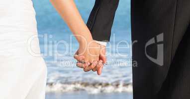 Bride and groom lower bodies holding hands against blurry beach shore