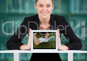 Business woman with tablet showing book on grass against blurry bookshelf with teal overlay