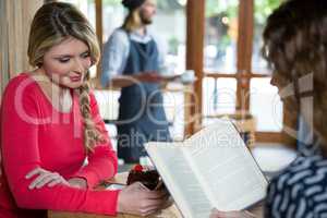 Woman using phone while friend reading book in coffee shop