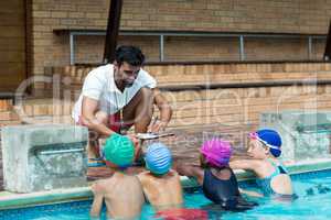 Instructor writing on clipboard while explaining little swimmers at poolside