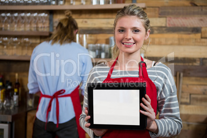 Portrait of smiling young woman showing digital tablet in background at coffee shop