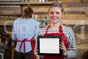Portrait of smiling young woman showing digital tablet in background at coffee shop