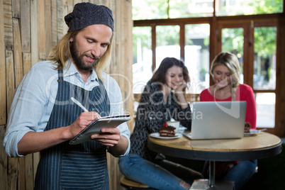 Male barista writing orders with female customers in background