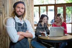 Confident male barista with female customers in background at cafe