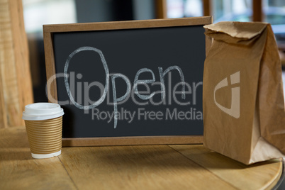 Open signboard with disposable coffee cup and paper bag on table