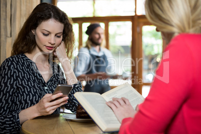 Young women using phone and reading book in coffee shop