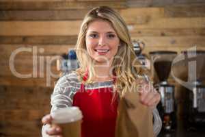 Smiling female barista holding coffee cup and paper bag in cafeteria