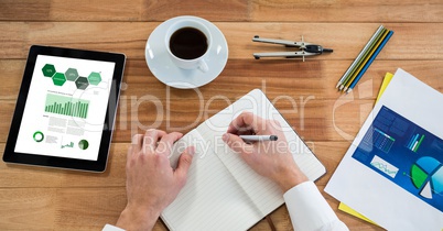 Cropped hand of person writing on diary by graphs in digital tablet at table in office