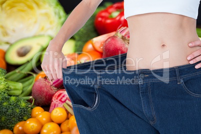 Midsection of woman wearing loose jeans with fruits and vegetable in background representing weight