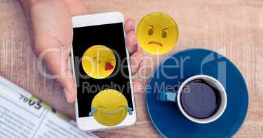 Digitally generated image of emojis flying over hands using smart phone by coffee cup at table