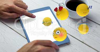 Digital composite image of hand using tablet PC with emojis flying over table