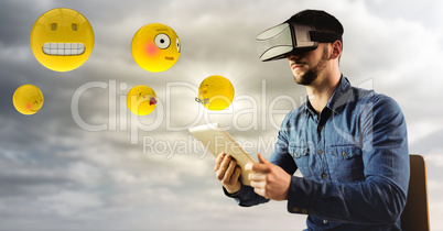 Man in VR using tablet with emojis and flare against cloudy sky