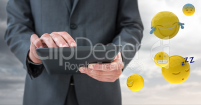 Business man mid section with tablet next to emojis and flare against cloudy sky