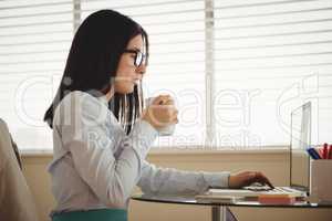 Side view of woman drinking coffee while sitting by laptop