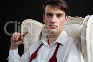 Androgynous man sitting on chair with cigar against black background