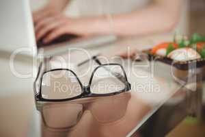 Eyeglasses on table against woman using laptop