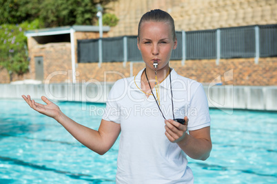 Female coach with stopwatch blowing whistle at poolside