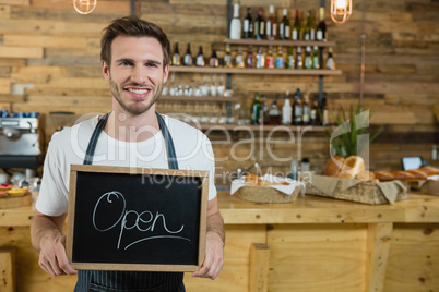 Portrait of smiling waiter standing with open signboard at counter