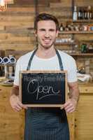 Portrait of smiling waiter standing with open signboard at counter