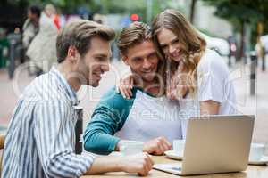 Cheerful couple with friend using laptop while sitting at sidewalk cafe