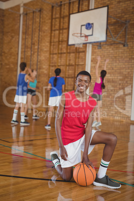 Smiling school boy kneeling with a basketball while team playing in background