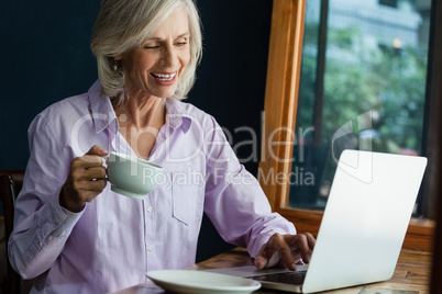 Smiling senior woman drinking coffee while working on laptop