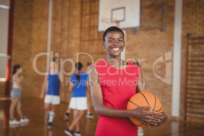 Smiling school boy holding a basketball while team playing in background
