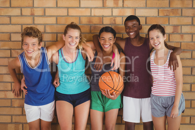 Happy high school kids with arms around leaning against the wall in basketball court
