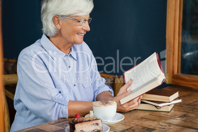 Smiling senior woman reading book while sitting by coffee at table