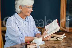 Smiling senior woman reading book while sitting by coffee at table