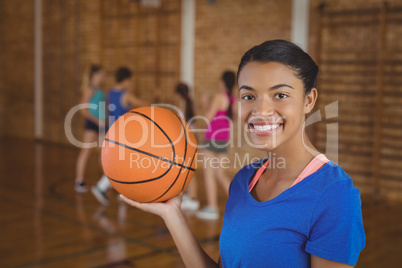 Smiling high school girl holding a basketball while team playing in background