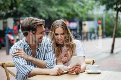 Cheerful couple holding mobile phone while sitting at sidewalk cafe