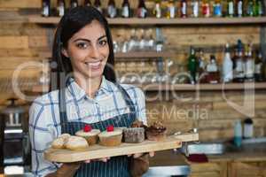 Portrait of smiling waitress holding cupcakes in wooden tray at counter