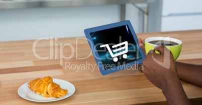 Cropped image of person holding digital tablet with shopping cart icon on screen while having coffee
